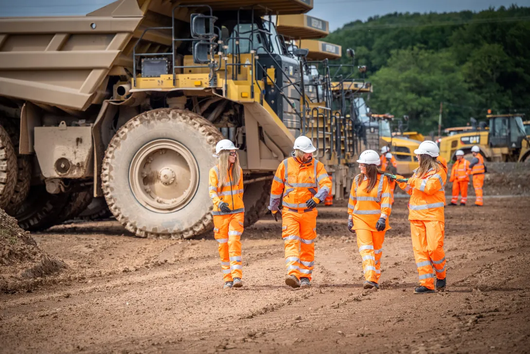 Four Aggregate Industries colleagues in high visibility workwear walk through Bardon Hill Quarry with heavy duty large dumper trucks in the background.